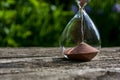 Hourglass with pouring sand against a background of blurred greenery. The bottom of the glass flask