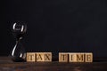 Hourglass or sandglass on cube wooden block with alphabets TAX TIME on wood table in the dark room.Tax Time Concept