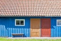 House of blue wooden planks, red roof, two colorful doors and small windows