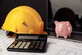 House building costs concept. Safety helmet and piggy bank with blueprint, calculator and laptop on table Royalty Free Stock Photo