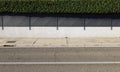 House exterior enclosure wall consisting of white plaster, iron fence and evergreen hedge above. Concrete sidewalk and paved road