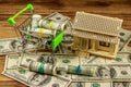 House model and small shopping cart with rolls of dollars on pile of  U.S. one hundred dollar bills on wooden background Royalty Free Stock Photo