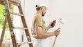 House painter man with mobile phone and paint roller, works the white home wall, a wooden ladder and green window as a background Royalty Free Stock Photo