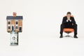 House on top of roll of bills with pensive businessman on chair representing expensive real estate