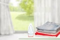 Household laundry ironing. Close-up of a red electrical iron and a stack of ironed clothes on white board over blurred curtains Royalty Free Stock Photo