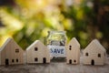 Houses model on wooden table with blurred coins in jar and sunlight bokeh background.