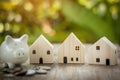 Houses model on wooden table. Blurred coins and piggy bank with sunlight bokeh background.