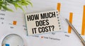 How Much Does it Cost text on a card against the background of a blank notebook on a desk, a business concept Royalty Free Stock Photo