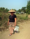 A Shan woman wearing a traditional straw hat walking on unpaved road with a plastic bag in her hand