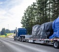 Huge blue classic big rig semi truck transporting covered heavy commercial cargo on step down semi trailer running on the wide Royalty Free Stock Photo