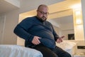 Huge man suffering from extra weight touching stomach sitting on bed in bedroom.