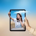 A human hand holding tablet with a screen view of an Asian girl in a hat and a suitcase taking a selfie with hills and a rock Royalty Free Stock Photo