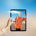 A human hand holding tablet with a screen view of an Asian girl in a scarf with a backpack standing with hills and a rock cliff Royalty Free Stock Photo