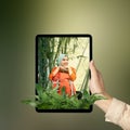 A human hand holding tablet with a screen view of an Asian girl in a scarf sitting with a backpack in the rainforest with trees Royalty Free Stock Photo