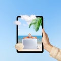 A human hand holding tablet with a screen view of an Asian girl in a scarf sitting with a suitcase on the beach with an ocean view Royalty Free Stock Photo