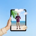 A human hand holding tablet with a screen view of an Asian man in a hat with a backpack standing on the beach with an ocean view Royalty Free Stock Photo