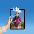 A human hand holding tablet with a screen view of an Asian man in a hat with a backpack and a suitcase standing with a mountain Royalty Free Stock Photo