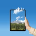 Human hand holding tablet with a screen view of the trees with mountain view and blue sky Royalty Free Stock Photo