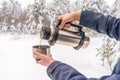 Human hands holding thermos with hot tea or coffee against landscape with snow covered forest. Man pouring drink into mug in Royalty Free Stock Photo