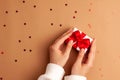 Human hands in white sweater puts a white paper gift with a red satin ribbon bow on right on brown background with red stars Royalty Free Stock Photo