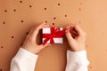 Human hands in white sweater untied a white paper gift with a red satin ribbon bow on brown background with red stars shapes. Royalty Free Stock Photo