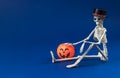 Human skeleton in black hat sits on blue background with basket in shape of pumpkin Jack-o-Lantern. Concept of Halloween