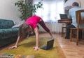 A mature woman performs yoga asana while a man works at a computer at home.