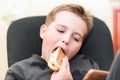 A Hungry boy eating a hot dog at home sit while playing with the smartphone.Close-up