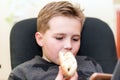 A Hungry boy eating a hot dog at home sit while playing with the smartphone.Close-up