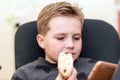 A Hungry boy eating a hot dog at home sit while playing with the smartphone.Close-up