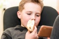 A Hungry boy eating a hot dog at home sit while playing with the smartphone.Close-up