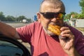 Hungry Cacasiam senior driver wearing sunglasses eating patty near his car standing on Ukrainian road