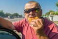 Hungry Caucasian senior driver in sunglasses eating junk food  fried patty near his car while doing short stop on Ukrainian road Royalty Free Stock Photo