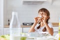 Hungry charming dark haired little girl biting and enjoying croissant, keeping eyes closed, sitting at table in kitchen, tasting