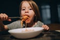 Hungry little girl appetite eating the bolognese pasta reeling up it on the fork and putting to mouth in the low light kitchen