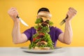 Hungry man with cutlery and huge burger at white table on yellow background