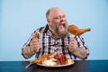 Hungry man with overweight eats chicken leg at table with fat food on blue background