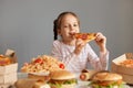 Hungry optimistic attractive little girl with braids sitting at table with junk food isolated over gray background biting big Royalty Free Stock Photo