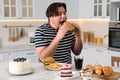 Hungry overweight man eating tasty burger at table in kitchen