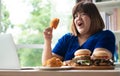 Hungry overweight woman holding Fried Chicken, hamburger on a wooden plate and Pizza on table, During work from home, gain weight