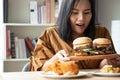 Hungry overweight woman holding hamburger on a wooden plate, Fried Chicken and Pizza on table, During work from home, gain weight
