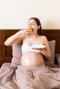 Hungry pregnant woman relaxing in bed is eating greedily a piece of cake and has a dirty mouth. Expecting mother can`t stop eatin Royalty Free Stock Photo