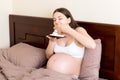 Hungry pregnant woman relaxing in bed is eating greedily a piece of cake and has a dirty mouth. Expecting mother can`t stop eatin Royalty Free Stock Photo