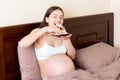 Hungry pregnant woman relaxing in bed is eating greedily a piece of cake and has a dirty mouth. Expecting mother can`t stop eatin Royalty Free Stock Photo