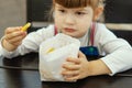 A hungry, pretty little girl holds a paper bag of French fries in her hand at a fast food cafe. Portrait of a happy child sitting