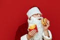 Hungry Santa Claus isolated on red background with burger and french fries in hands. Man in santa costume eating burger. Isolated Royalty Free Stock Photo