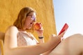 Hungry teenager with glasses biting an apple in the terrace with orange wall, sitting and studying in the smart phone. Blond short