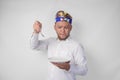 Hungry young Balinese man in white shirt and traditional headdress holding an empty plate with copy space and spoon while making a Royalty Free Stock Photo