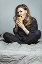 Hungry young woman in pajamas eating burger in bed. Beautiful blonde. Fast food and snacks. Gray background. Vertical Royalty Free Stock Photo