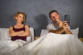 Husband or boyfriend using mobile phone in bed and suspicious frustrated wife or girlfriend feeling upset suspecting betrayal and